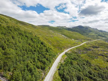 Drone fotoğrafı. Güneşli bir yaz gününde kümülüs bulutlarıyla mavi gökyüzünün altında asfalt karayolu olan Norveç dağ vadisinin en üst manzarası..