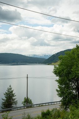 Norveç Doğal Manzarası. Büyük fiyort dağlarıyla çevrili mavi sularla çevrili bir gölün yanında bir otoyol var..