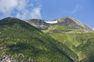 Yeşil geçitten, kayalık derin vadiye, keskin uçurumlar, siluetler, mavi gökyüzündeki bulutların altındaki karlı dağlara doğru giden hava manzaralı bir yol. Uzaktaki dev kar şatosuyla dramatik katmanlı manzara.