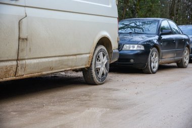 Riga, Letonya - 25 Aralık 2024: Kirli beyaz bir minibüs ve kırsal bir alanda çamurlu, asfaltsız bir yolda park edilmiş siyah bir sedan. Araçlar tozla kaplı, bu da off-road ya da sert hava koşullarını gösteriyor. Arkaplanda ağaç var