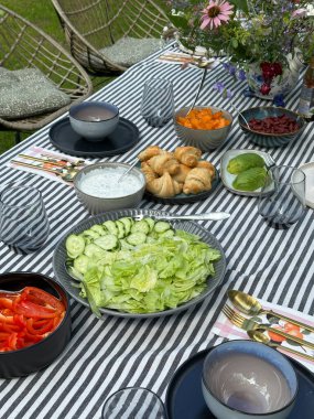 Ev yapımı ekmek, kruvasan, fitaki peyniri, bıldırcın yumurtası, domates, karabuğday ve ton balığıyla dolu bir çiftlik evinde yemek masası. Üzerinde çizgili bir masa örtüsü ve vazoda çiçekler var..