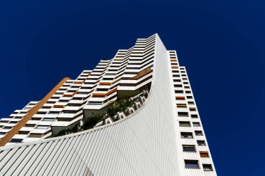 Açık mavi gökyüzünün altında kıvrımlı ön cepheli ve yeşil terasları olan modern yüksek binalar.