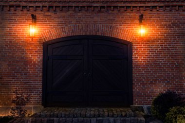 Ön taraftaki tuğla bina ve eski ahşap kapı iki lambayla aydınlatılır.