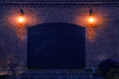 Ön taraftaki tuğla bina ve eski ahşap kapı iki lambayla aydınlatılır.