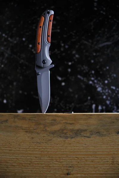 Folding knife for survival stuck wooden beam, on dark background