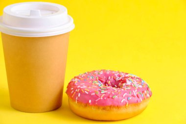 Disposable coffee cup and doughnut with pink icing yellow background