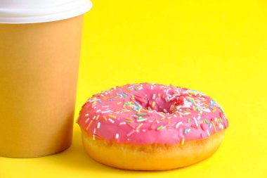 Disposable coffee cup and doughnut with pink icing yellow background