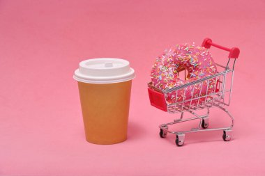 Pink donut in shopping cart and disposable coffee cup on pink background