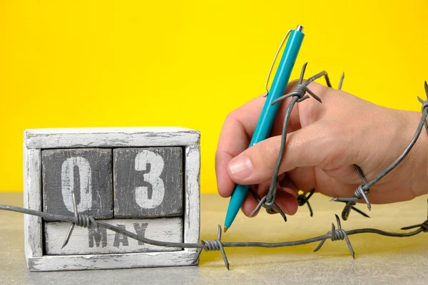 Hand wrapped barbed wire holds ballpoint pen on yellow background.On calendar May 3.Concept for Press Freedom Day