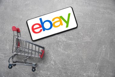 Kostanay, Kazakistan, 31 Ağustos 2023. ebay logolu akıllı telefon, alışveriş sepeti gri beton arka plan. İnternetten alışveriş kavramı.