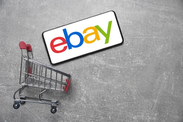 Kostanay, Kazakistan, 31 Ağustos 2023. ebay logolu akıllı telefon, alışveriş sepeti gri beton arka plan. İnternetten alışveriş kavramı.