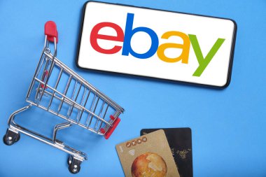Kostanay, Kazakistan, 31 Ağustos 2023. ebay logolu akıllı telefon, alışveriş sepeti ve banka kartları mavi arka plan. İnternetten alışveriş kavramı.