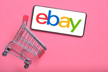 Kostanay, Kazakistan, 31 Ağustos 2023 ebay logolu akıllı telefon, alışveriş sepeti pembe arka plan. Çevrimiçi alışveriş kavramı.
