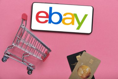 Kostanay, Kazakistan, 31 Ağustos 2023. ebay logolu akıllı telefon, alışveriş sepeti ve banka kartları pembe arka plan. Çevrimiçi alışveriş kavramı