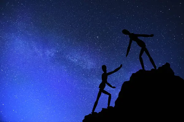 Two silhouettes mannequin climb mountain, one helping other, extending helping hand. Against background night sky. Concept of help and success in teamwork