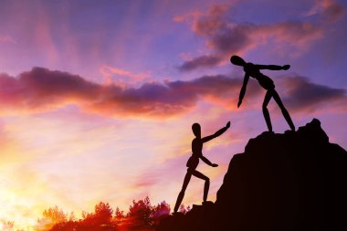İki siluet manken dağa tırmanıyor, biri diğerine yardım ediyor, yardım elini uzatıyor. Arka plandaki gökyüzüne, günbatımına ve bulutlara karşı. Takım çalışmasında yardım ve başarı kavramı