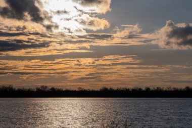 Gölün üzerinde gün batımı önplanda ağaçlar ve bulutlu gökyüzü