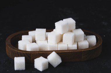 Tahta bir tabakta beyaz şeker küpleri. Siyah arka planda tahta kasede küp şekerler..