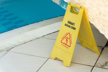 Yüzme havuzu tarafından yerleştirilen sarı uyarı levhası, insanları fayanslı zeminde kaygan zemine karşı uyararak, eğlence alanlarında güvenlik ve ihtiyatı teşvik ediyor.