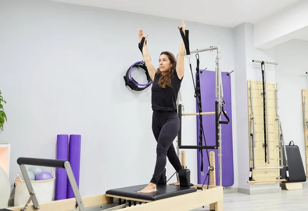 Full body of female in activewear standing on Pilates reformer and stretching raised arms with resistance bands during training at gym