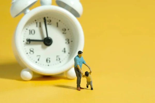Miniature people toy figure photography. Time for children concept. A father and son standing in front of white clock. Isolated yellow background. Image photo