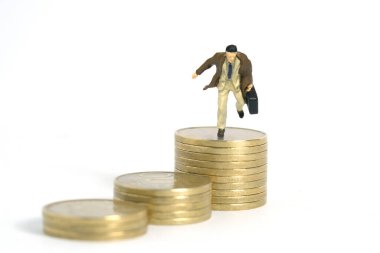 Minyatür minyatür insanlar oyuncak figürlü fotoğrafçılık. Altın madeni para yığınının üstüne atlayan palto giyen bir işadamı. Beyaz arka planda izole edilmiş. Resim fotoğrafı.