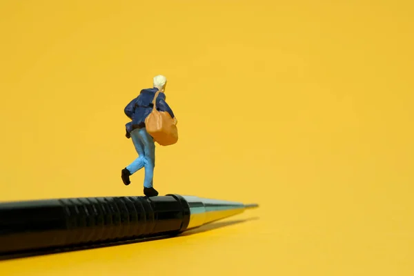 Miniature tiny people toy figure photography. A college student running above pencil carrying bag. Isolated on yellow background. Image photo