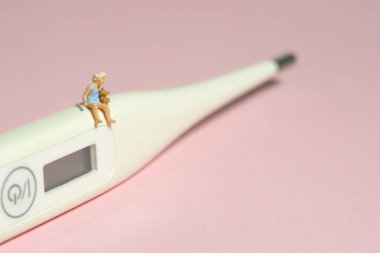 Minyatür minyatür insanlar oyuncak figürlü fotoğrafçılık. Beyaz termometrenin üzerinde oturan küçük bir kız oyuncak ayı bebeği tutuyor. Pembe arka planda izole edilmiş. Resim fotoğrafı.
