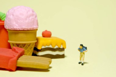 Minyatür insanlar oyuncak fotoğrafçılık. Sağlıklı beslenme ve öğle yemeği arası konsepti. Çocuk bir anaokulu öğrencisi aperatif ve pastanın yanında duruyor. Sarı arka planda izole edilmiş. Resim fotoğrafı.