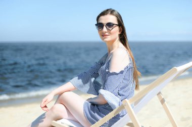 Mutlu genç esmer kadın, okyanus sahilinde ahşap bir iskemlede dinlenirken gülümsüyor ve gözlük takıyor. Keyifli tatil konsepti.