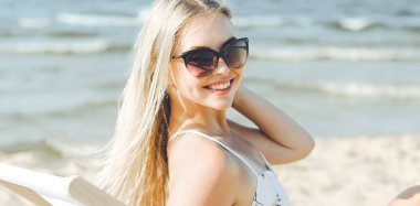 Mutlu sarışın kadın güneş gözlüğü takıyor ve okyanus sahilinde tahta bir iskemlede dinleniyor..