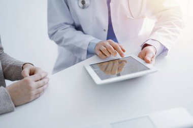 Doktor ve hasta klinikteki masada oturuyor. Tablet bilgisayar kullanan kadın doktorların ellerine odaklan, kapat. Tıp ve sağlık hizmetleri kavramı.