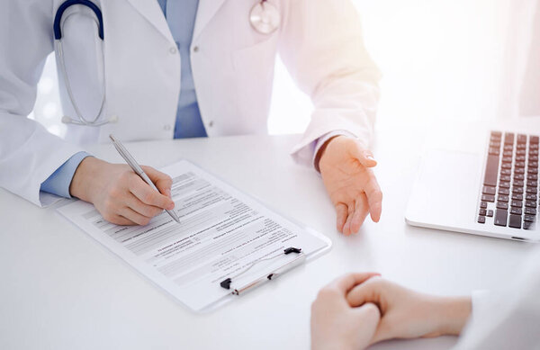 Doctor and patient sitting opposite each other at the desk in clinic. The focus is on female physicians hands filling up the medication history record form or checklist, close up. Medicine concept.
