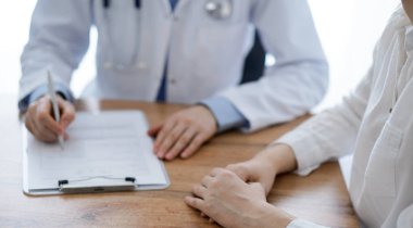 Doktor ve hasta klinikteki ahşap masada oturuyorlar. Kadın doktorların ellerinin bir kayıt formunu doldurmasına odaklanıyoruz. Tıp konsepti.