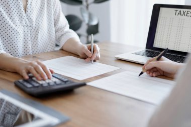 Ofisteki ahşap masada vergileri sayarken iki muhasebeci dizüstü bilgisayar ve hesap makinesi kullanıyor. İş denetimi ve finans alanında takım çalışması.