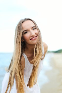 Okyanus sahilinde kameraya bakarak mutlu, gülümseyen bir kadın. Beyaz yaz elbisesi içinde çok kültürlü bir kadın modelin portresi açık havada tatil sırasında doğanın tadını çıkarıyor..