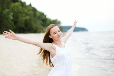 Okyanus sahilinde mutlu, mutlu ve mutlu bir kadın elleri açık duruyor. Beyaz yaz elbisesi içinde çok kültürlü bir kadın modelin portresi seyahat tatilinde doğanın tadını çıkarıyor.