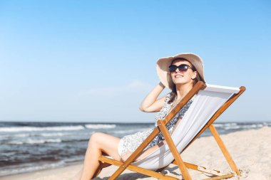 Mutlu esmer kadın güneş gözlüğü takıyor ve şapka takıyor. Okyanus sahilinde tahta bir iskemlede dinleniyor..