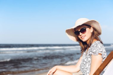 Mutlu esmer kadın güneş gözlüğü takıyor ve şapka takıyor. Okyanus sahilinde tahta bir iskemlede dinleniyor..