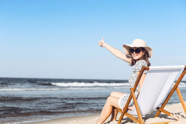 ハッピーブルネットの女性は 親指を示しながら海岸の木製のデッキの椅子に座っています — ストック写真
