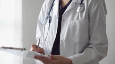Doktor kadın, tıbbi notlar yazarken beyaz bir not panosu kullanıyor. Hastanenin panorama penceresinin yanında duran bilinmeyen bir doktor. İlaç..