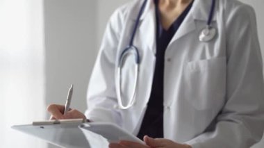 Doktor kadın, tıbbi notlar yazarken beyaz bir not panosu kullanıyor. Hastanenin panorama penceresinin yanında duran bilinmeyen bir doktor. İlaç..