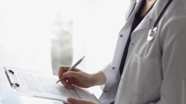 Doktor kadın, tıbbi notlar yazarken beyaz bir not panosu kullanıyor. Hastanenin panorama penceresinin yanında duran bilinmeyen bir doktor. İlaç..