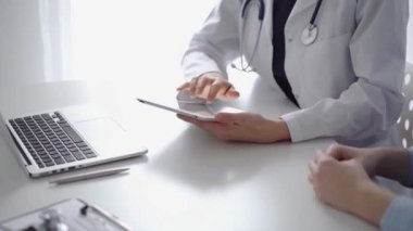 Doktor ve hasta klinikteki özel pencerenin yanındaki beyaz masada oturuyorlar. Beyaz önlük giyen kimliği belirsiz kadın doktor tıbbi kayıt formlarını doldurmak için tablet bilgisayar kullanıyor. Tıp konsepti.