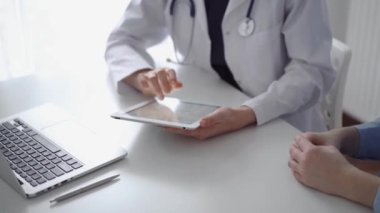 Doktor ve hasta klinikteki özel pencerenin yanındaki beyaz masada oturuyorlar. Beyaz önlük giyen kimliği belirsiz kadın doktor tıbbi kayıt formlarını doldurmak için tablet bilgisayar kullanıyor. Tıp konsepti.