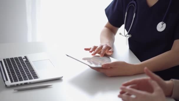 医生和病人坐在诊所亚麻窗附近的白桌前 不为人知的女医生用平板电脑填写病历表 医学概念 — 图库视频影像
