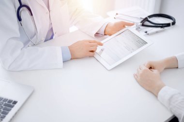 Doktor ve hasta klinikteki masada karşılıklı oturuyorlar. Kadın doktorların elleri tablet bilgisayarın dokunmatik dokunmatik pedini işaret ediyor, kapat. Tıp konsepti.