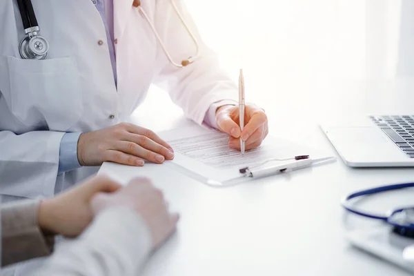 stock image Doctor and patient sitting at the table in clinic. The focus is on female physicians hands filling up the medication history record form or checklist, close up. Medicine concept.