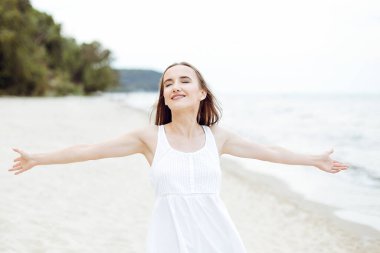 Okyanus sahilinde mutlu, mutlu ve mutlu bir kadın elleri açık duruyor. Beyaz yaz elbisesi içinde çok kültürlü bir kadın modelin portresi seyahat tatilinde doğanın tadını çıkarıyor.