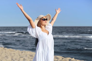 Mutlu sarışın kadın okyanus sahilinde beyaz elbiseli, güneş gözlüklü ve şapkalı el kaldırıyor..